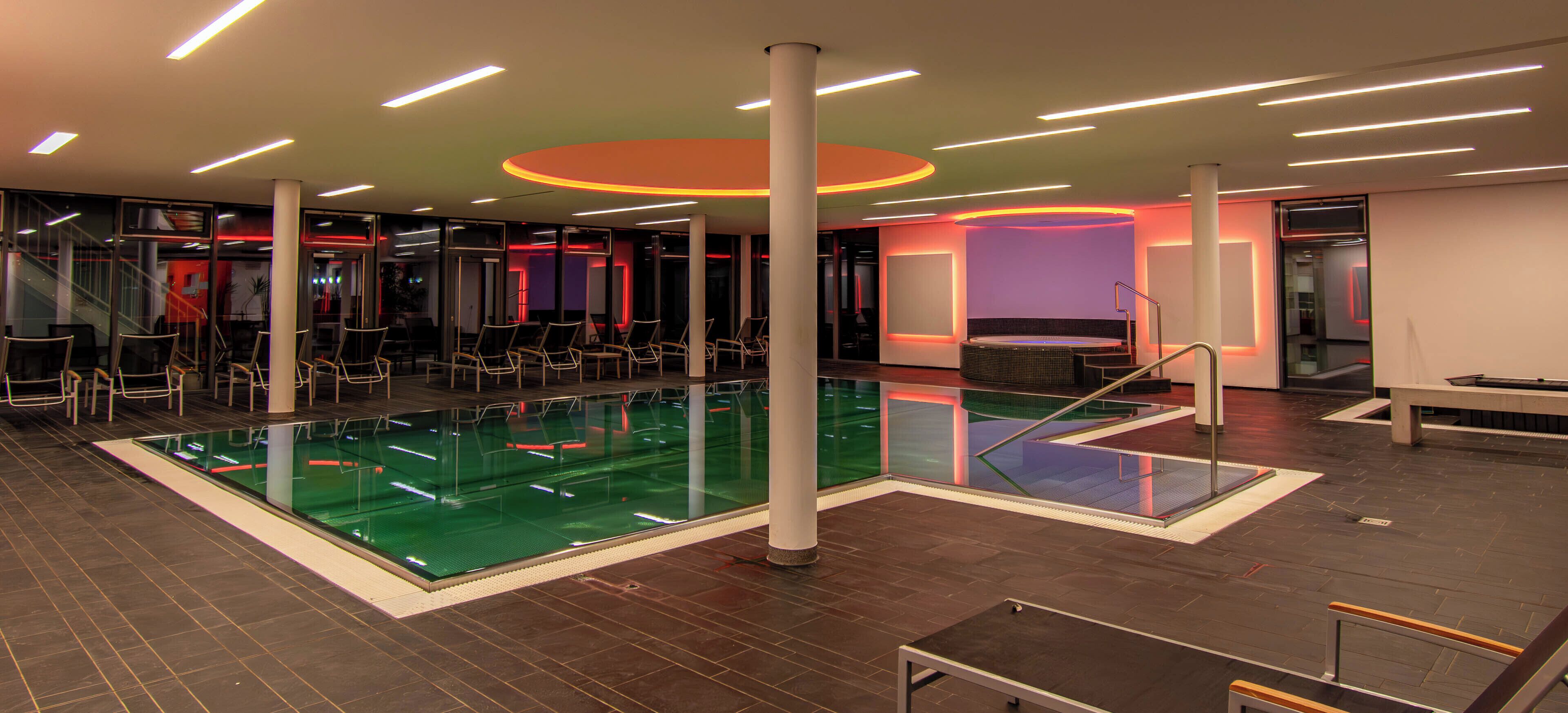 A modern indoor swimming pool with multiple lanes, surrounded by lounge chairs and a glass wall.