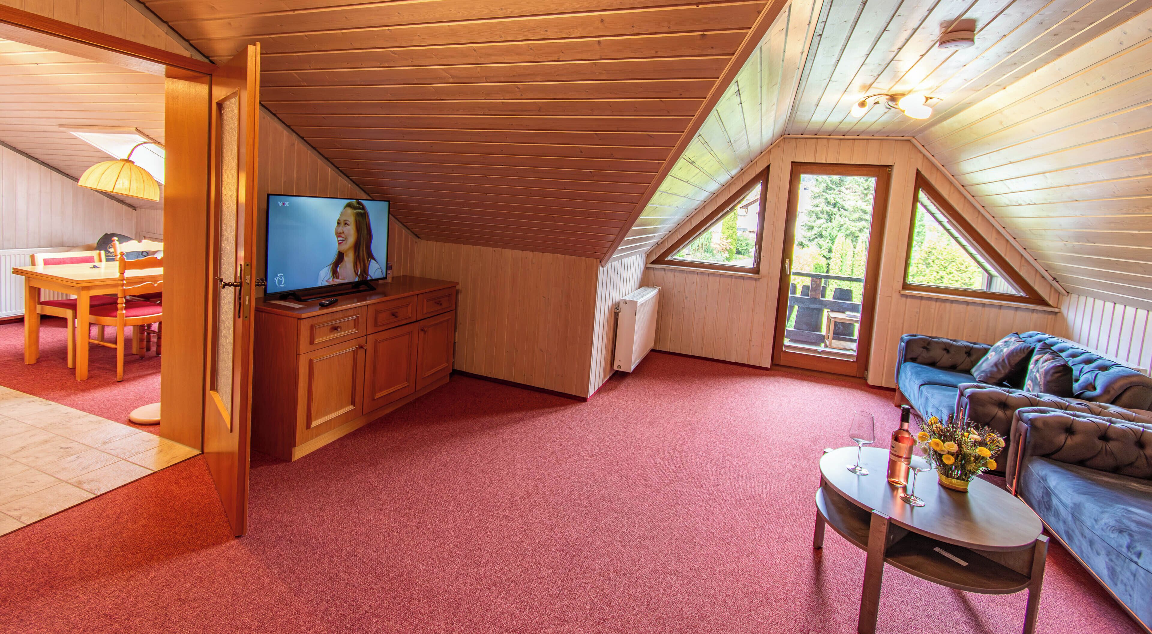 Ein gemütliches Dachzimmer mit Fernseher, Holzmöbeln, einem roten Teppich und Blick ins Freie.