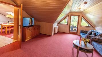 Ein gemütliches Dachzimmer mit Fernseher, Holzmöbeln, einem roten Teppich und Blick ins Freie.