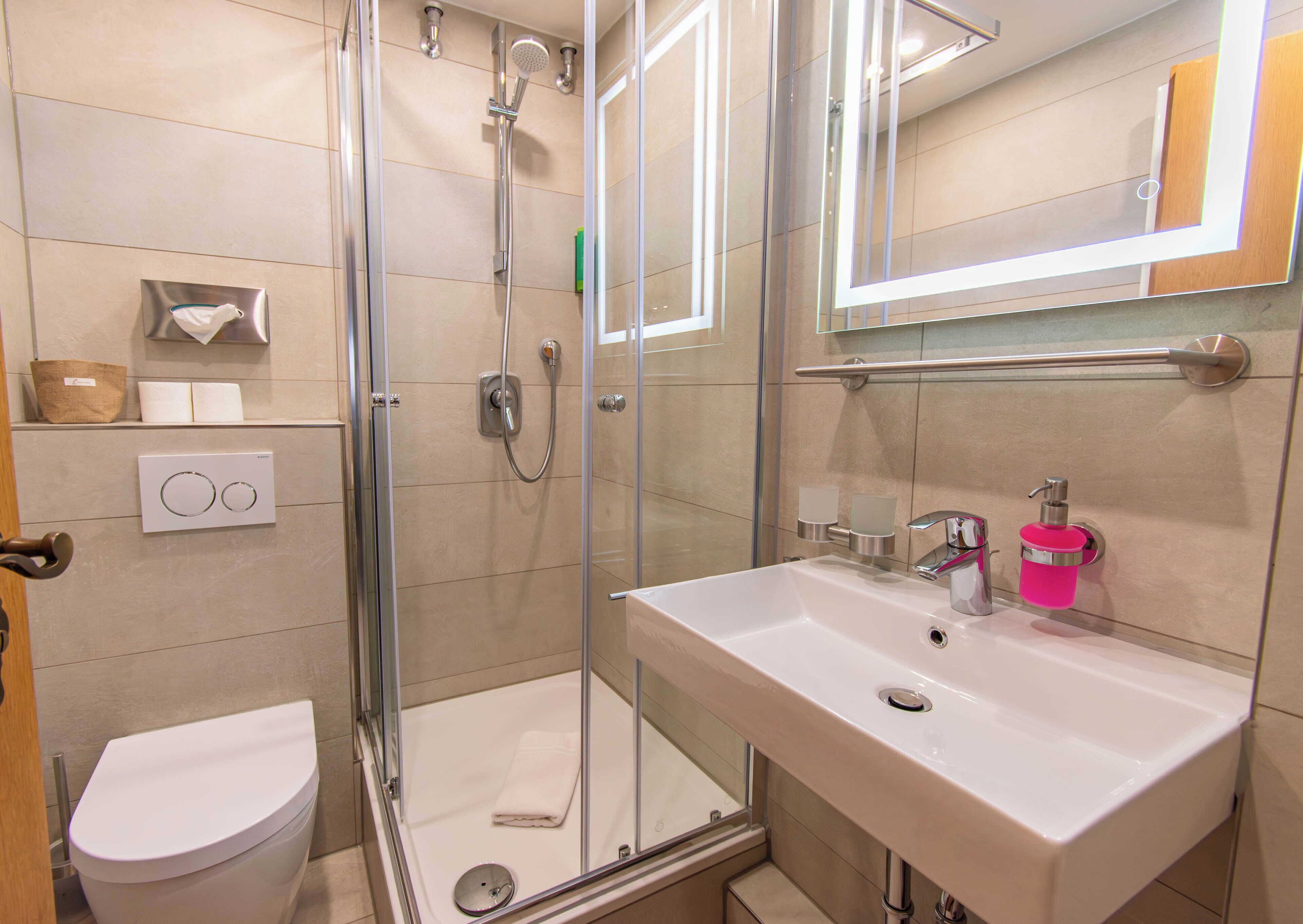 Une salle de bain moderne avec une douche à parois de verre, un lavabo blanc doté d’un distributeur de savon rose et des toilettes.