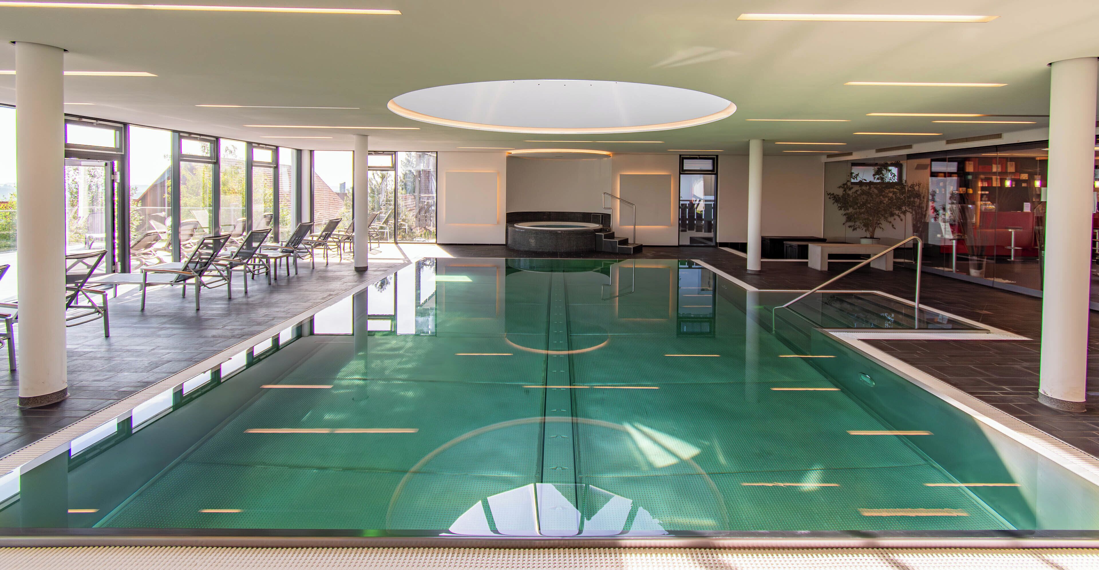 Piscina interior moderna com paredes de vidro transparente, área de estar iluminada pelo sol e uma banheira de hidromassagem central.