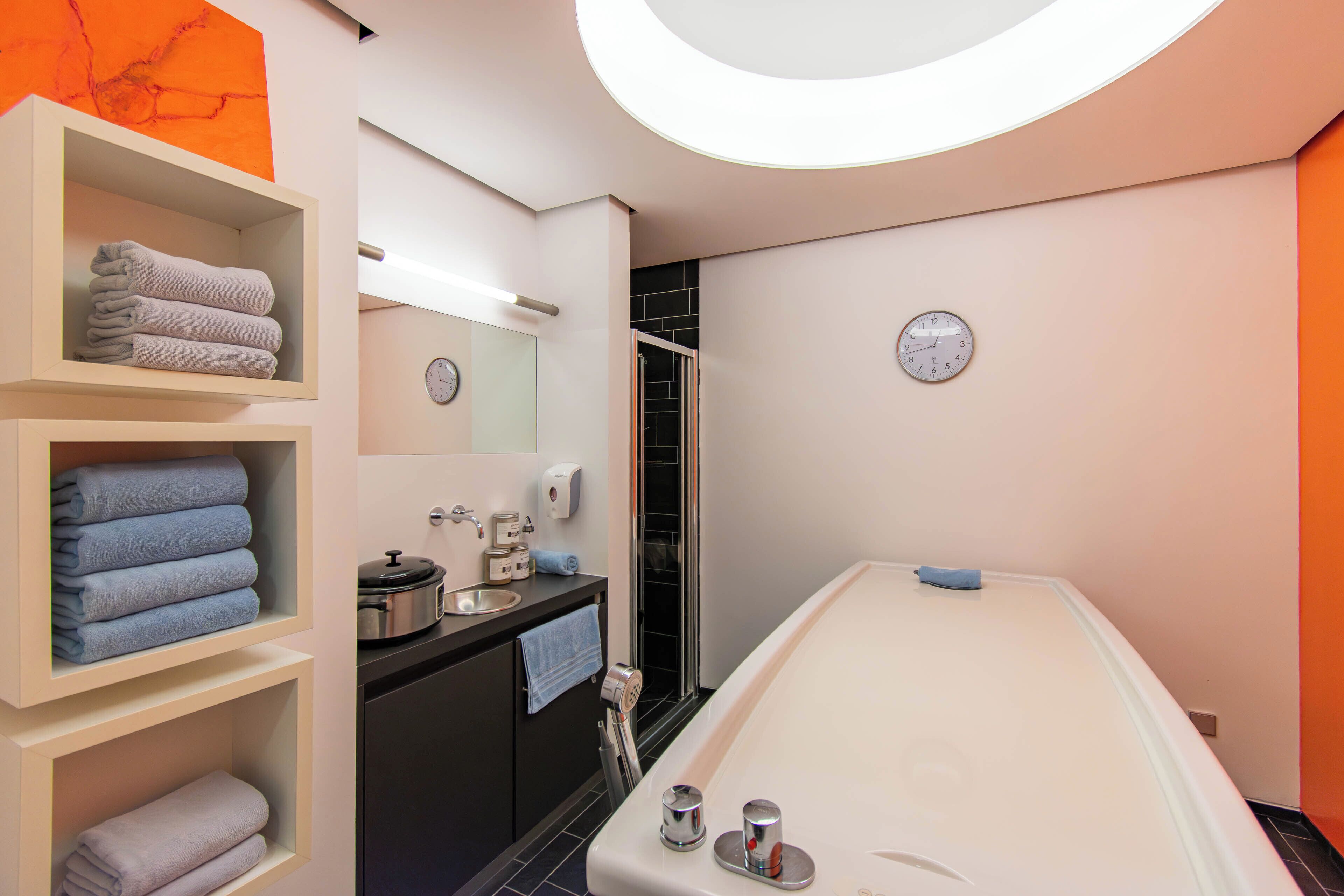 A modern bathroom with a bathtub, shelves with towels, a sink with a soap dispenser, and a clock on the wall.
