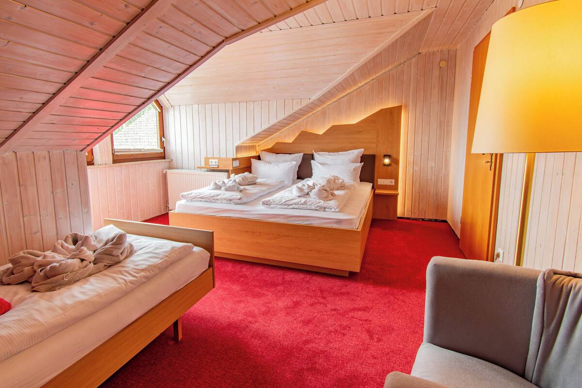 Quarto com teto inclinado de madeira, cama com lençóis brancos, tapete vermelho e um banco de madeira.