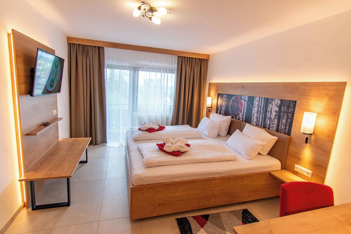 Quarto de hotel com cama de madeira, uma escrivaninha, televisão e uma janela grande com cortinas.