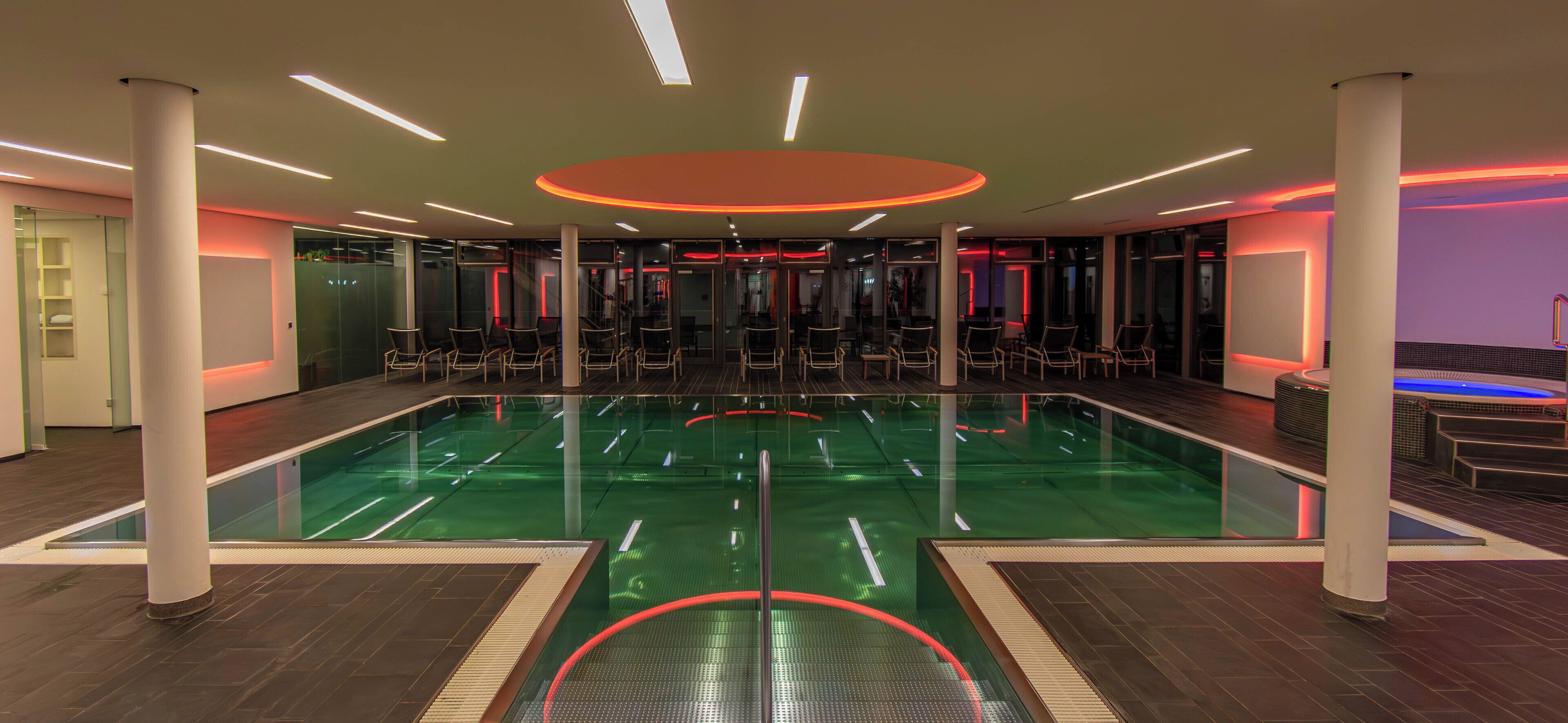 A modern indoor swimming pool with glass walls, seating areas, and a central circular light fixture.