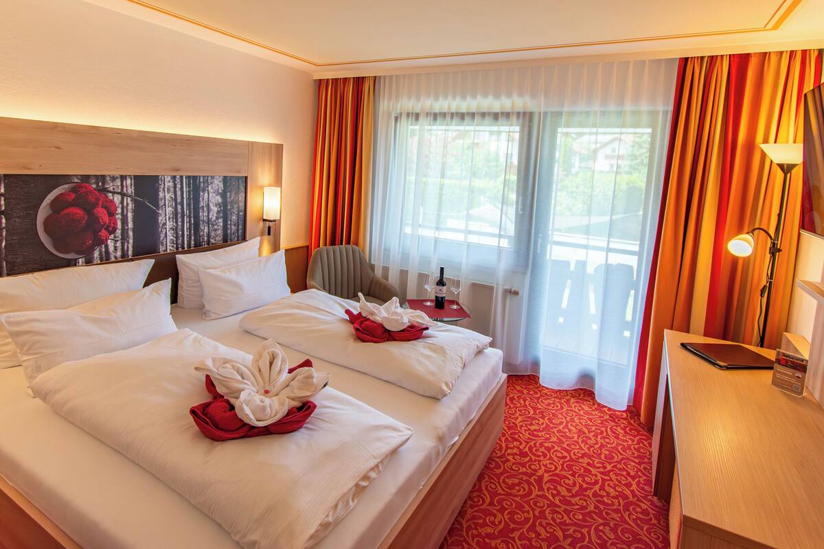 Quarto de hotel com uma cama grande, duas almofadas, uma cadeira, uma escrivaninha, um abajur e um quadro na parede.