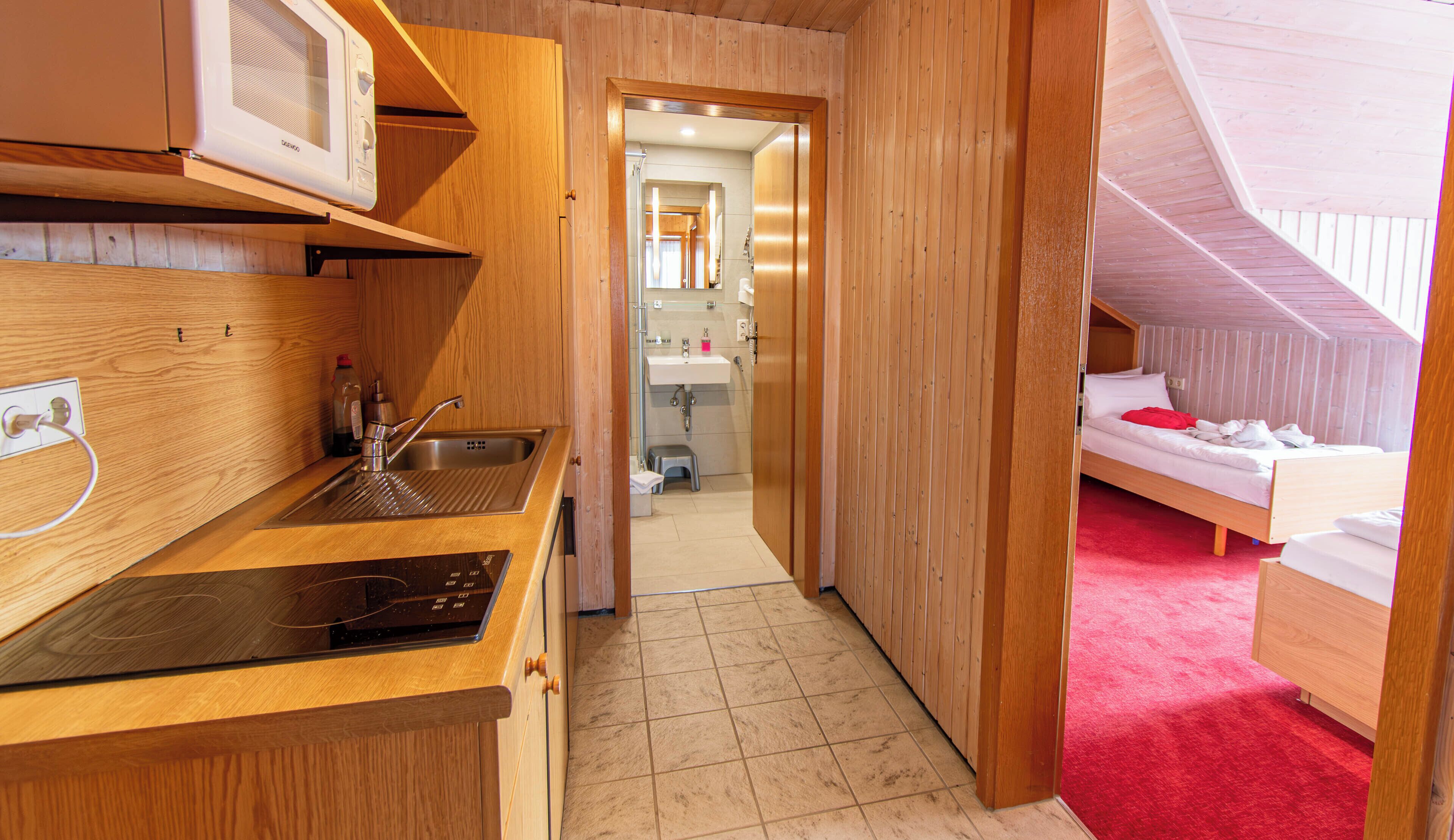 A compact, wooden-themed room with a kitchenette, a small bathroom, and a bed with a red accent.