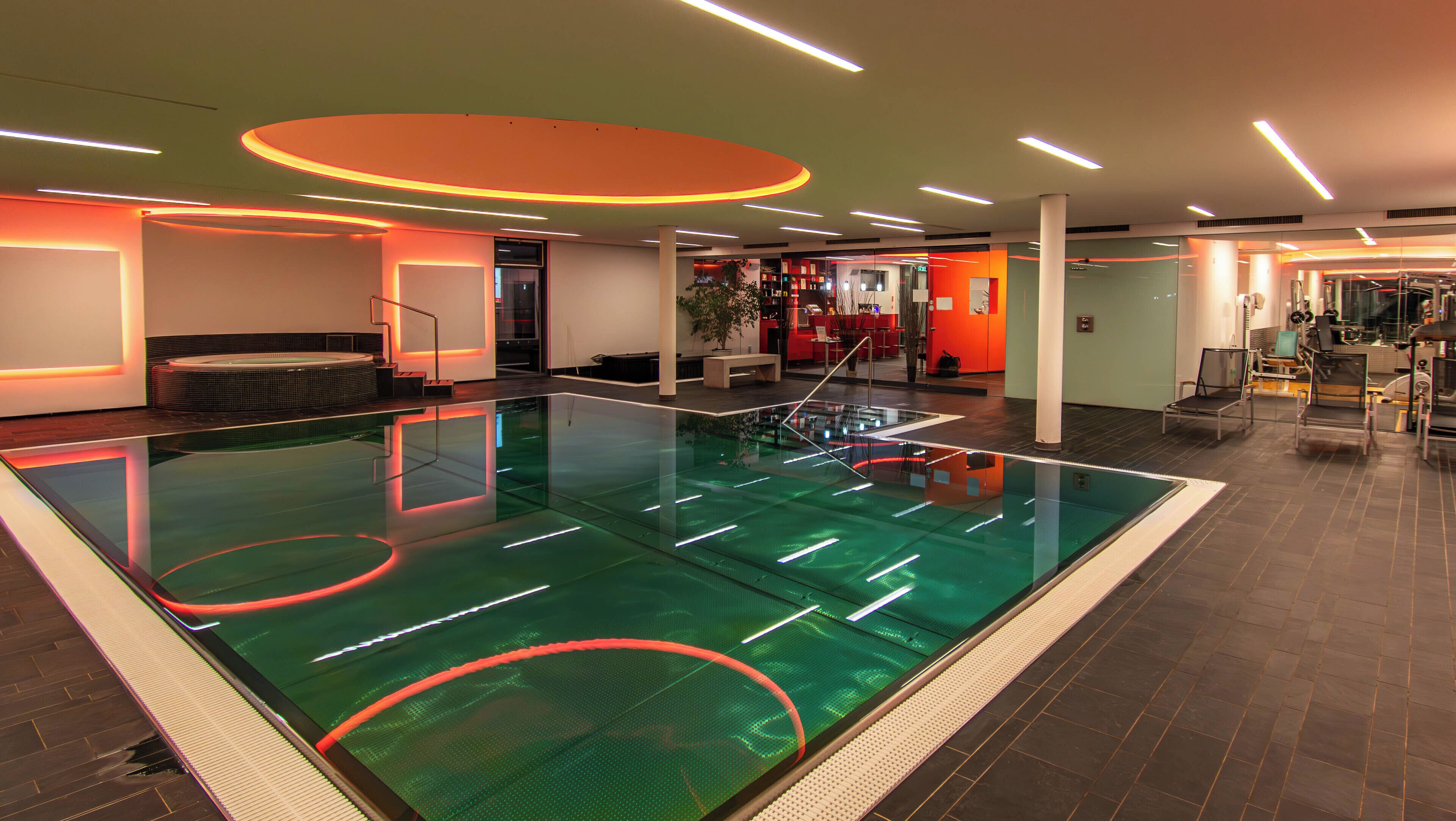 A modern indoor swimming pool with a glass floor, surrounded by a lounge area with seating and a reception desk.
