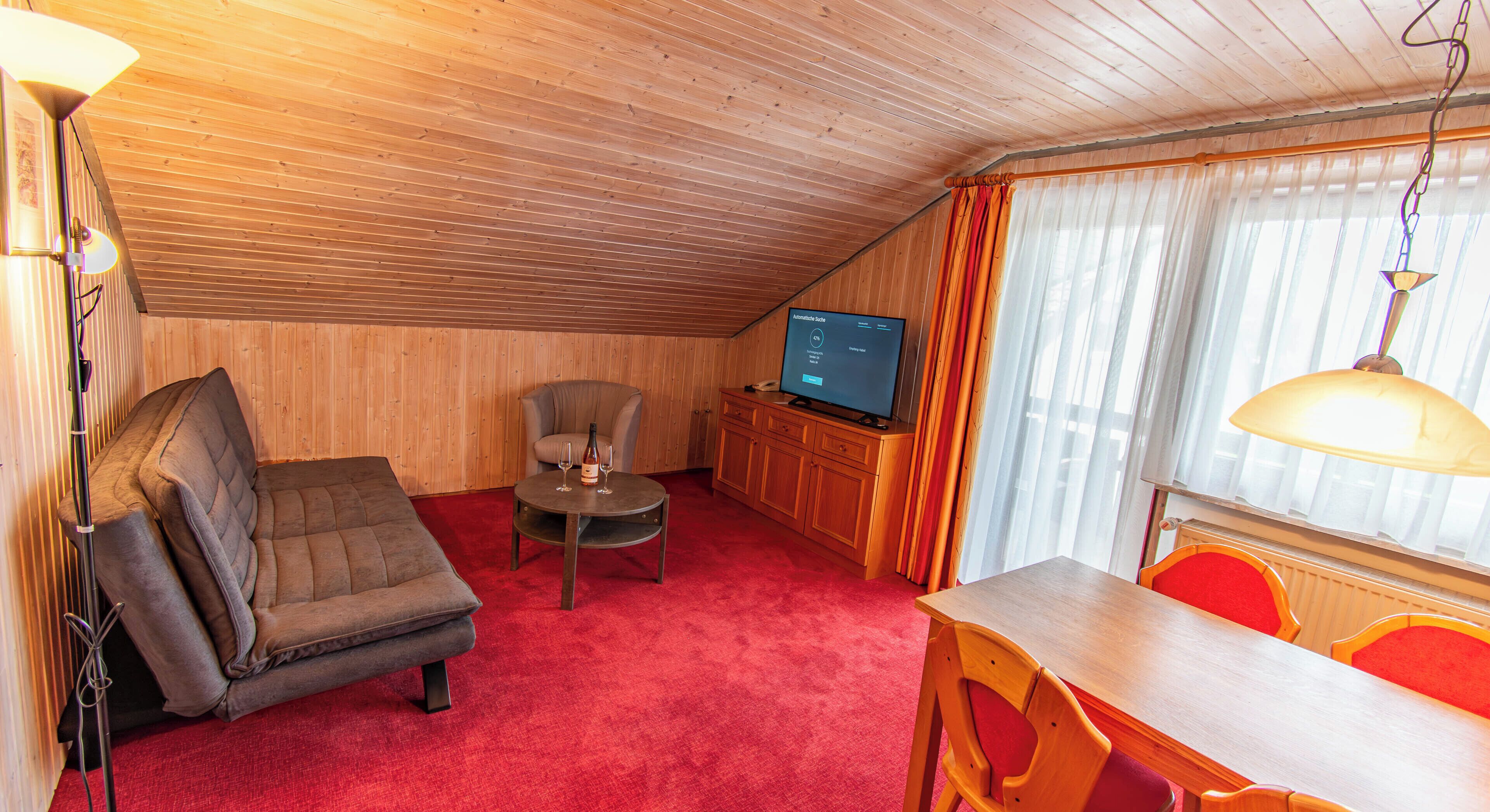 Een gezellige kamer met houten plafond, een rood tapijt, een bank, een eettafel met stoelen en een televisie.