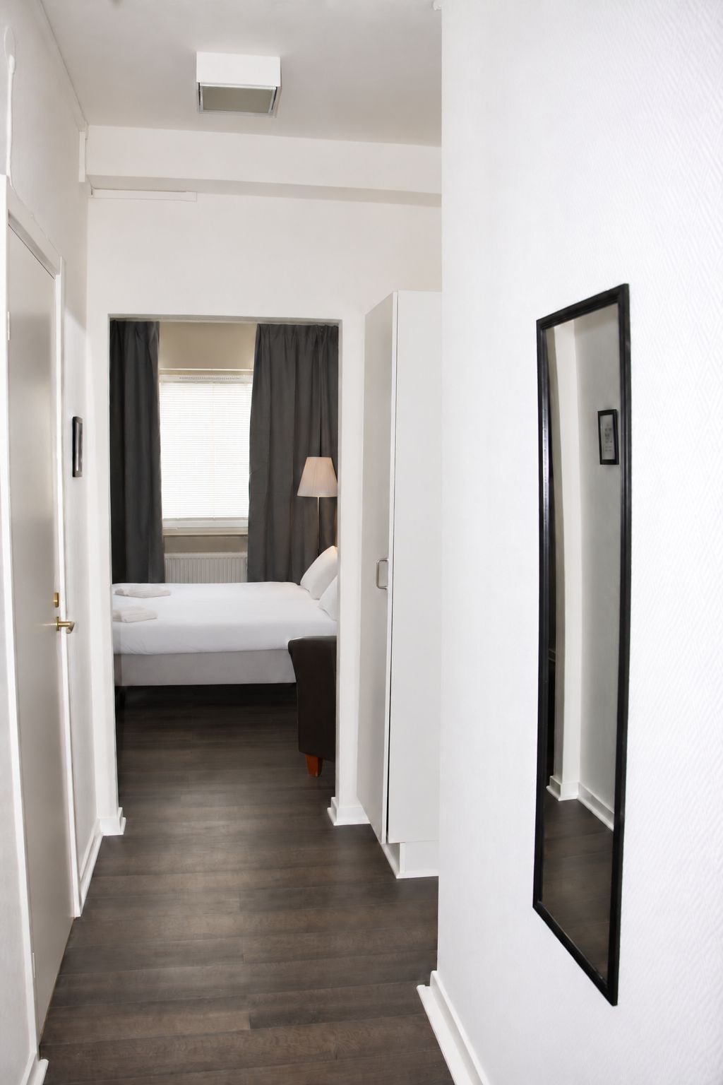 Ein Hotelzimmer mit Bett, Nachttischlampe, einem Fenster mit Vorhängen und einem Flur, der in ein weiteres Zimmer führt.