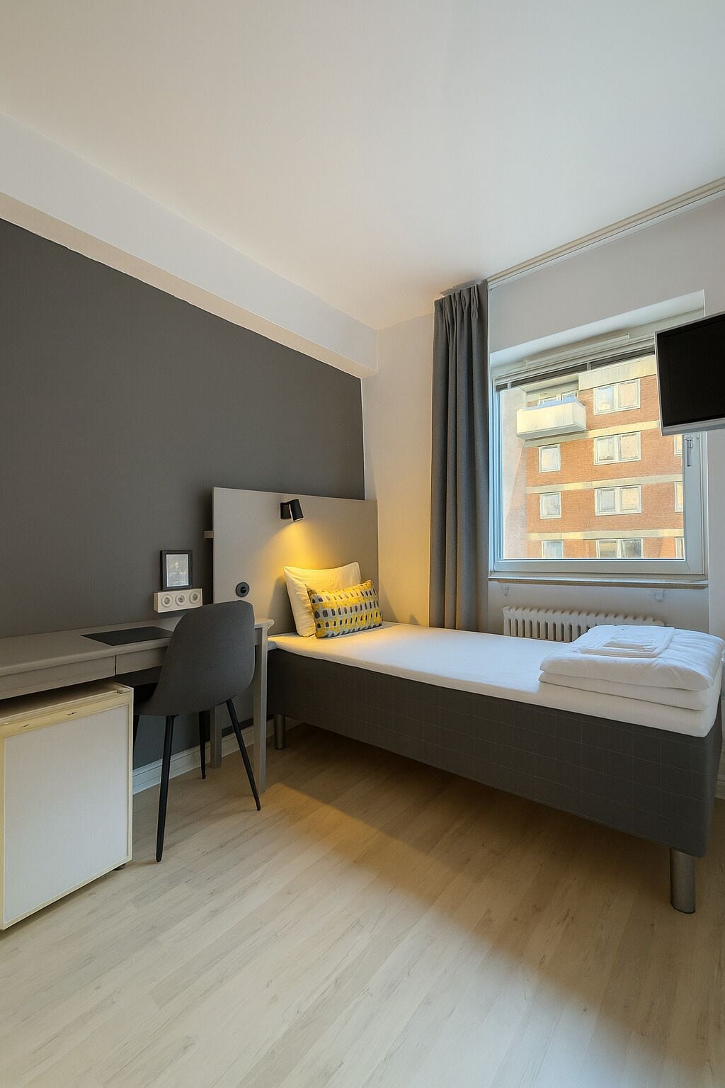 Quarto compacto com cama, escrivaninha, cadeira e janela com vista para um edifício.