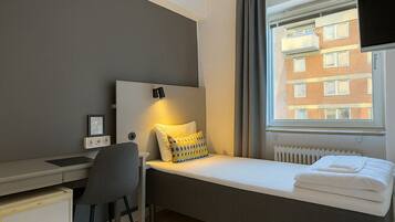 Quarto compacto com cama, escrivaninha, cadeira e janela com vista para um edifício.