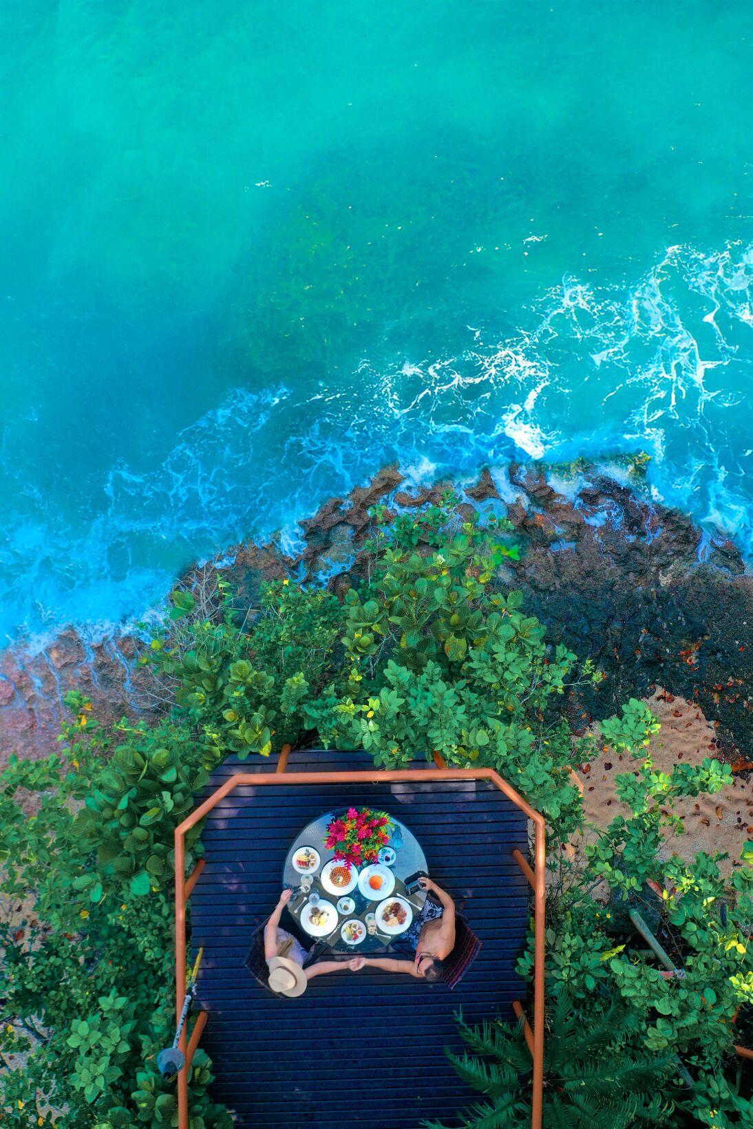 An aerial view of a dining area with a table set for two, overlooking a turquoise sea and rocky shoreline.