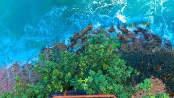 An aerial view of a dining area with a table set for two, overlooking a turquoise sea and rocky shoreline.