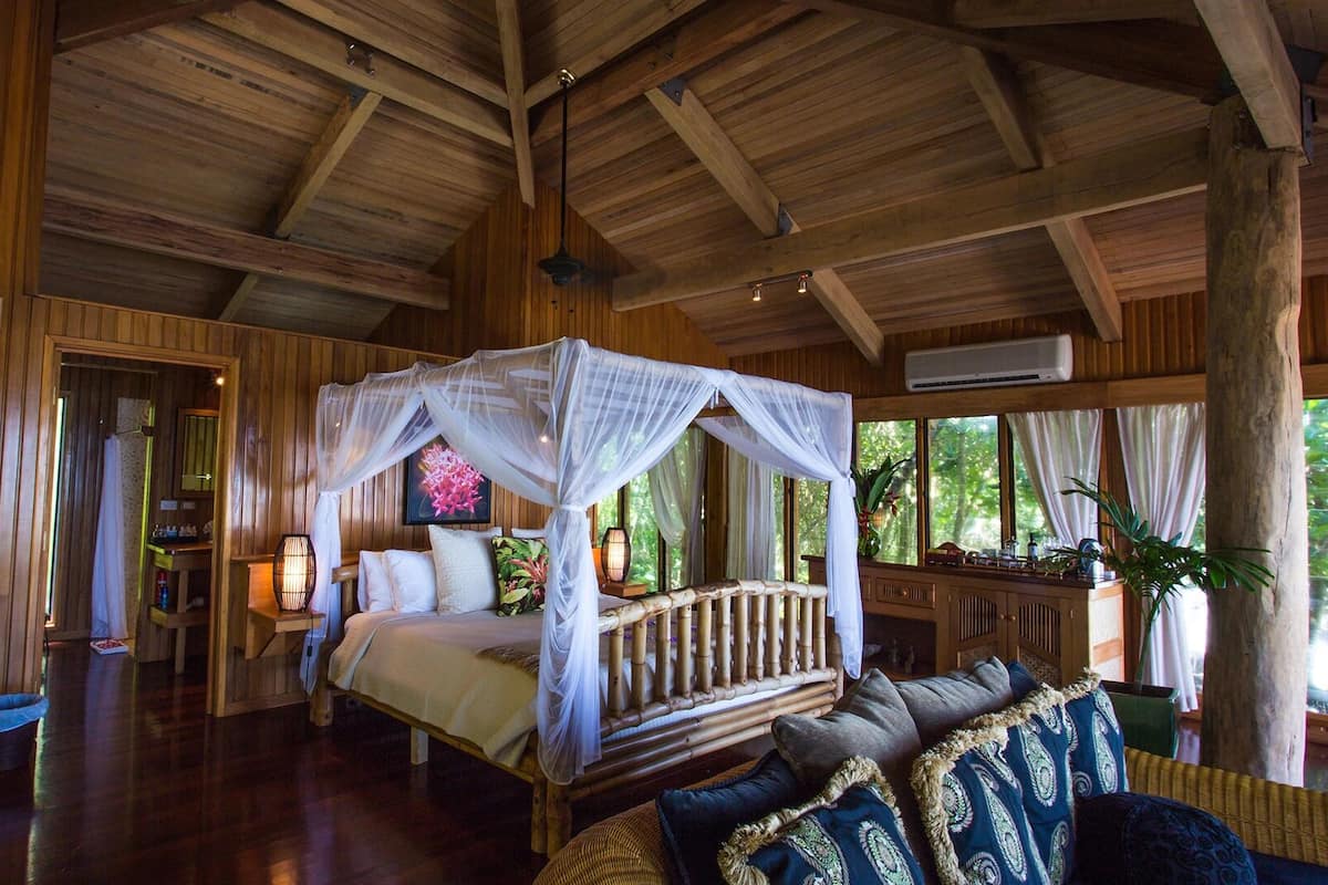Una habitación amplia con una cama grande, dosel y techo de madera. Se observa un sofá y una mesita con una lámpara.
