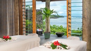 A room with two beds, a stone wall, a potted plant, and a view of the ocean.