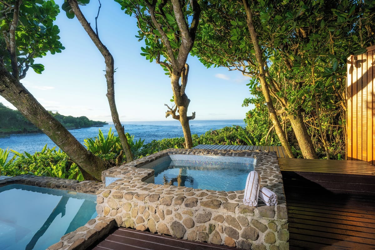 Un jacuzzi rodeado de muros de piedra, una terraza de madera y abundante vegetación con vista al mar.