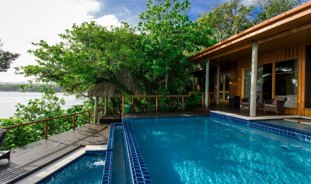 A swimming pool with a wooden deck and lounge area, surrounded by lush greenery and overlooking a body of water.