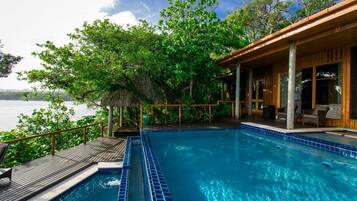 A swimming pool with a wooden deck and lounge area, surrounded by lush greenery and overlooking a body of water.
