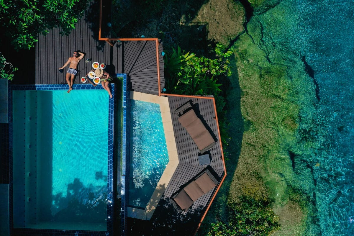 Vista aérea de una piscina con zona de solarium, rodeada de exuberante vegetación y agua azul cristalina.