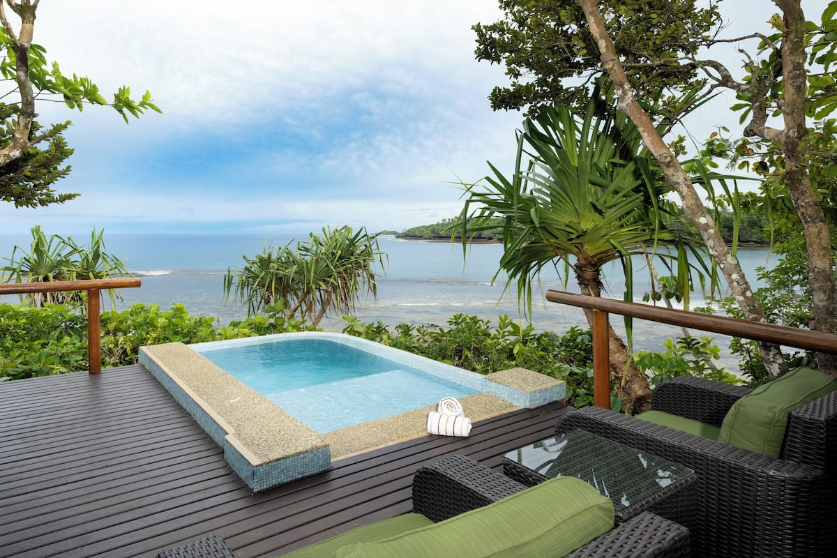 Zona de piscina con vista al mar, rodeada de vegetación y mobiliario de mimbre.
