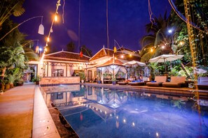 A pool area with lounge chairs and umbrellas, surrounded by potted plants and a building with a tiled roof.