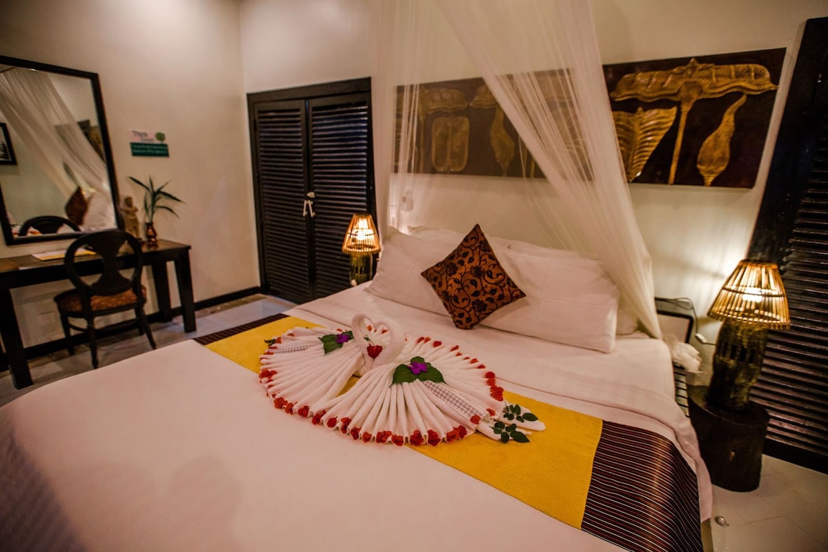 A hotel room with a bed, a desk, a chair, a mirror, and a nightstand with a lamp.