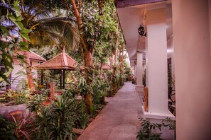 A pathway lined with tropical plants and palm trees, leading to a building with a tiled roof and white walls.