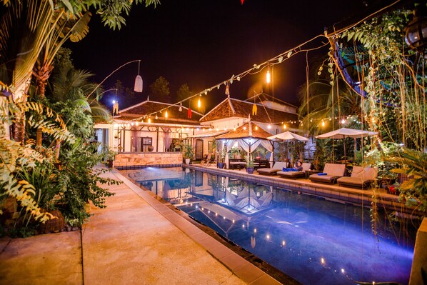 A pool area with lounge chairs and umbrellas, surrounded by greenery and string lights.