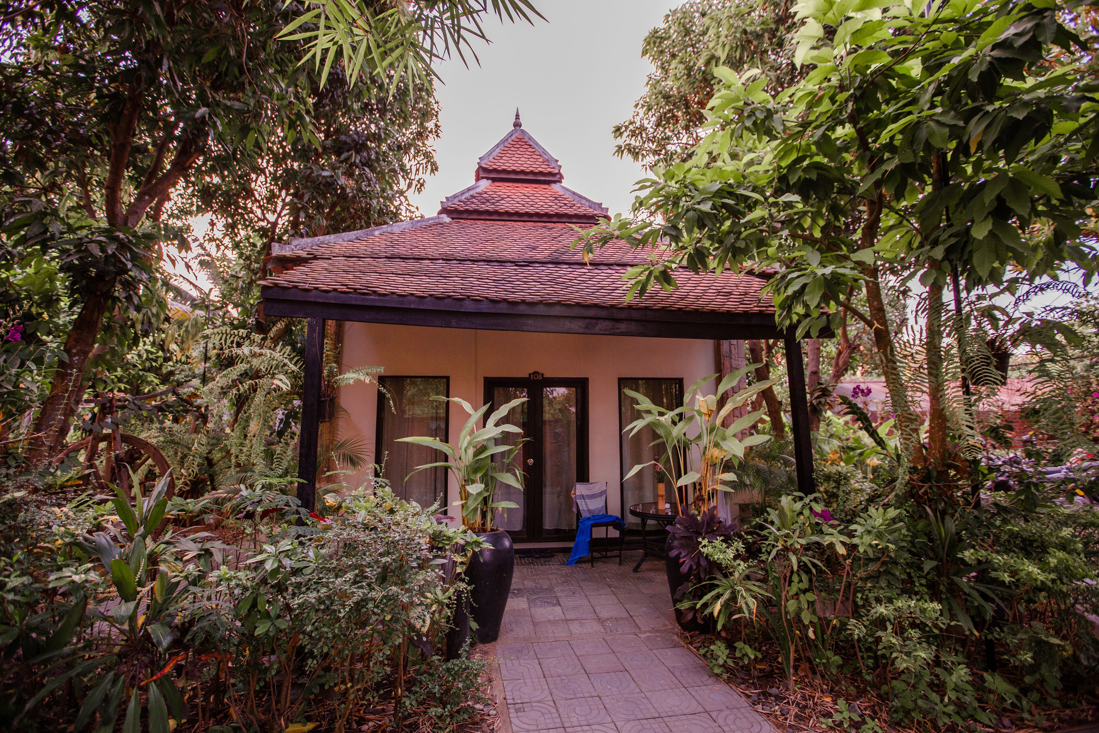 A traditional building with a tiled roof, surrounded by lush greenery and a paved pathway leading to a door.