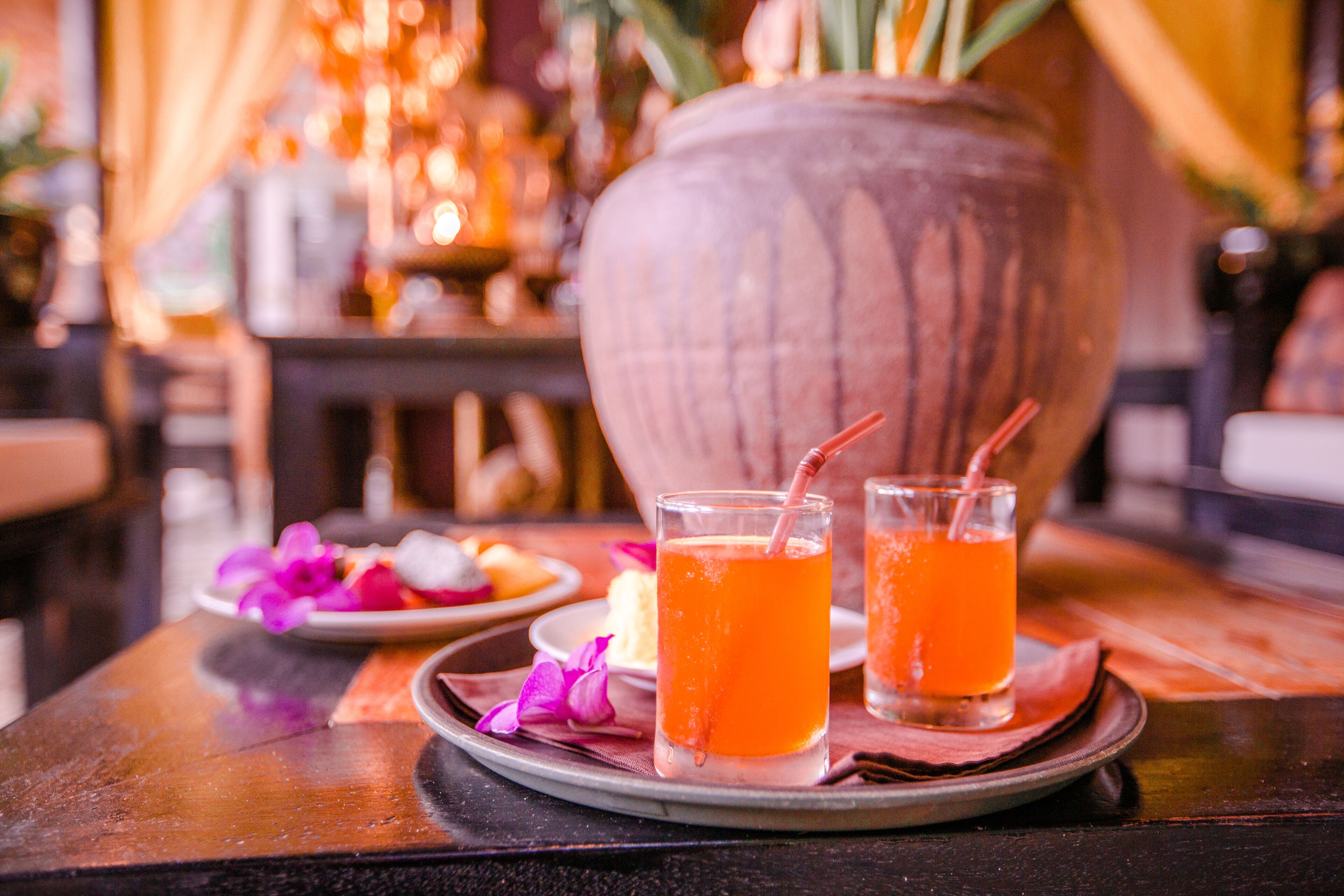 Two glasses of orange beverage on a tray with a plate of food and a decorative vase in the background.