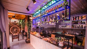 A bar area with shelves of bottles, a counter with a cash register, and a large wooden wheel in the background. - The Sanctuary Villa Battambang (Battambang)