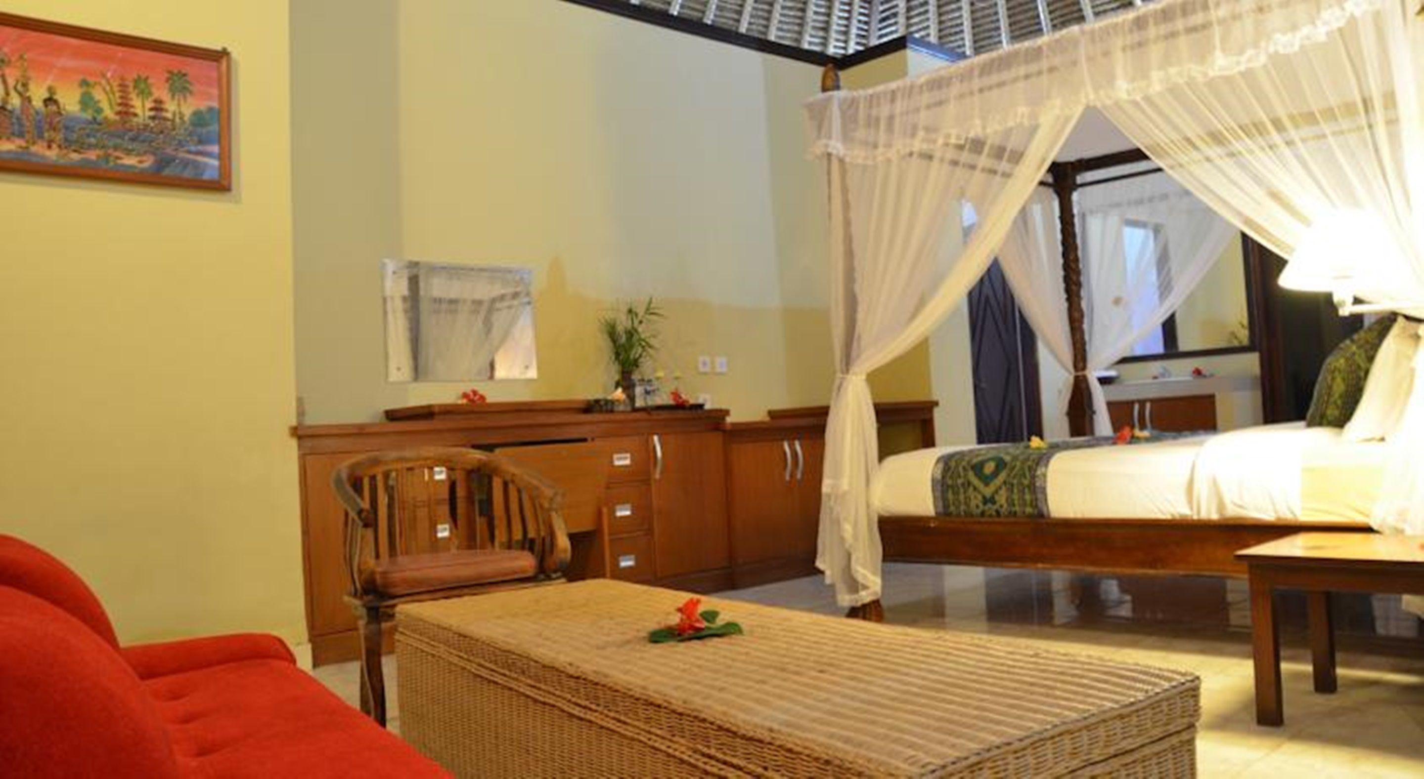 A bedroom with a canopy bed, a wooden cabinet, a wicker ottoman, and a painting on the wall.