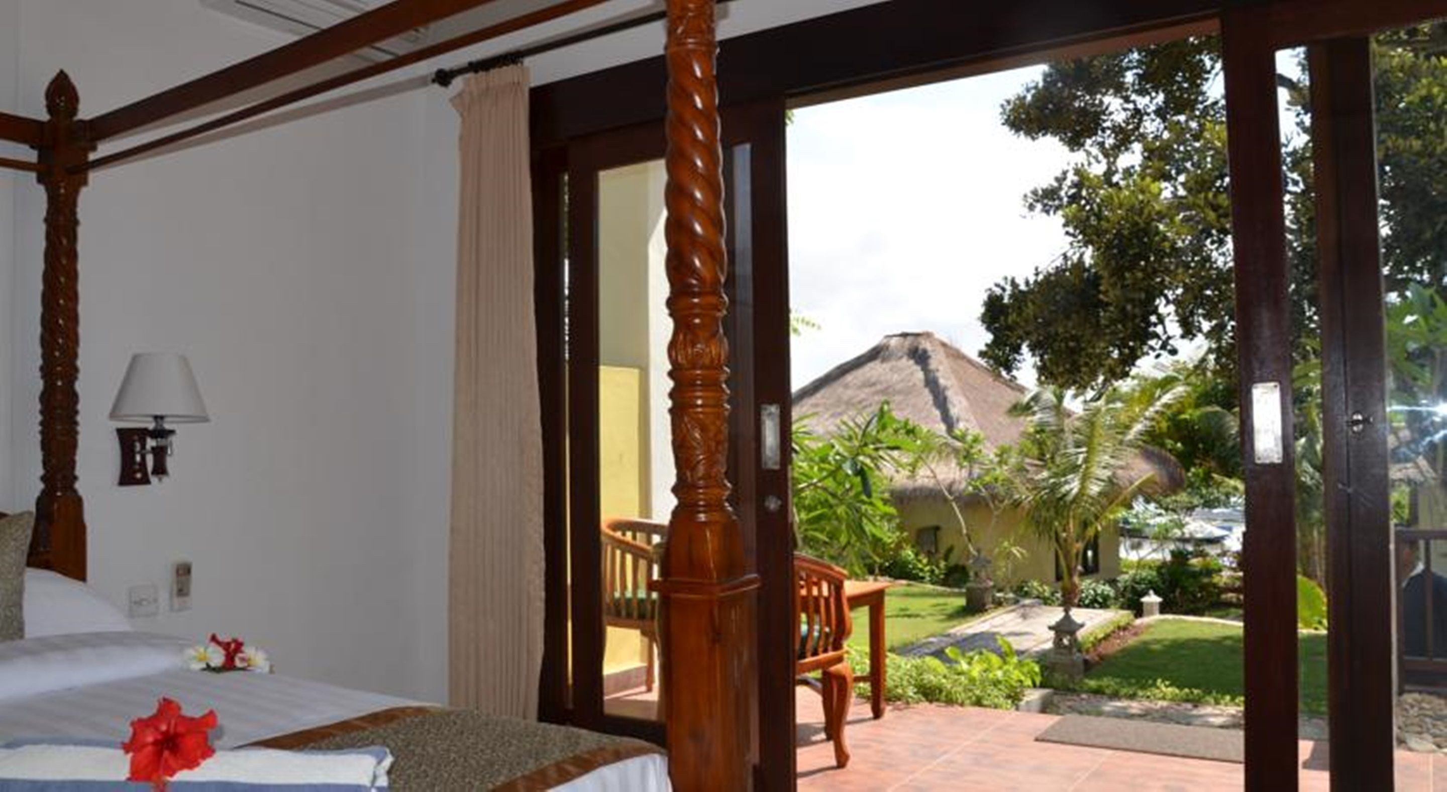 A bedroom with a large bed, a wooden chair, and a view of a thatched-roof building and greenery outside.