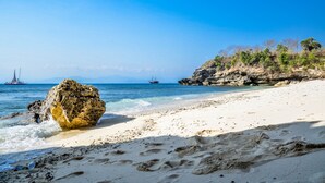 A sandy beach with clear blue water, a large rock in the foreground, and a rocky outcrop with sparse vegetation on the right.
