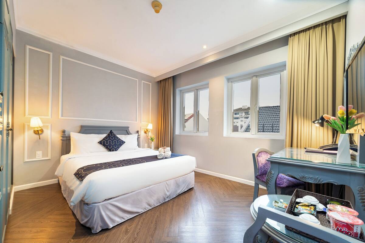 Ein Hotelzimmer mit einem großen Bett, Nachttischlampen, einem Schreibtisch mit Blumenvase und einem Fenster mit Vorhängen.