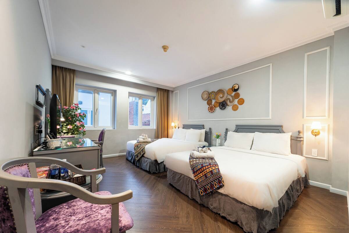 Ein Hotelzimmer mit zwei Betten, einem Schreibtisch und einem Stuhl. Es gibt Fenster mit Vorhängen, eine Wanduhr und eine dekorative Wandgestaltung.