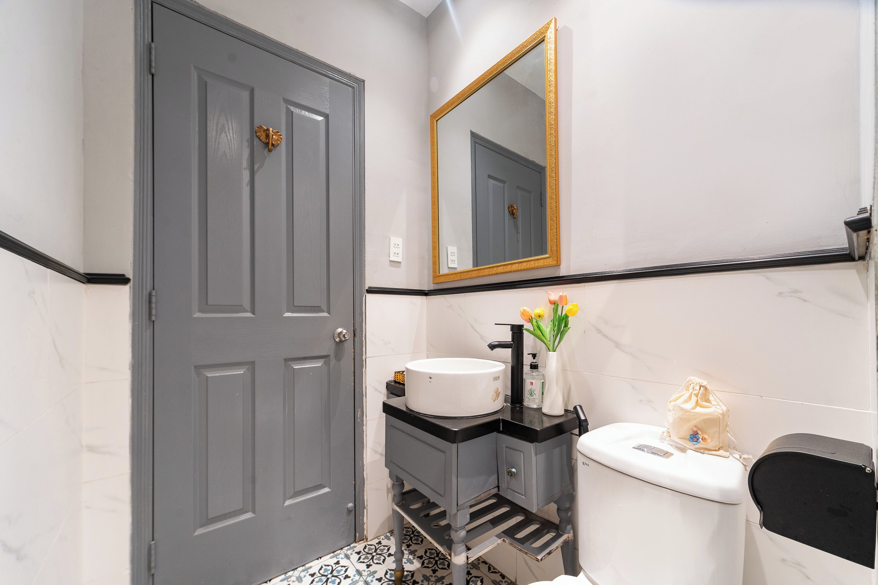 A bathroom with a grey door, a white sink with a black countertop, a mirror with a gold frame, and a toilet with a black lid.
