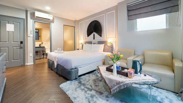 A hotel room with a bed, a sofa, a small table with a vase of flowers, and a window with a view. - Sabina Residence - Stellar Boutique Hotel (Ho Chi Minh City)