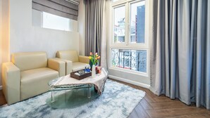 A modern living room with a beige sofa, a glass coffee table, and a view of the city through a large window with grey curtains. - Sabina Residence - Stellar Boutique Hotel (Ho Chi Minh City)