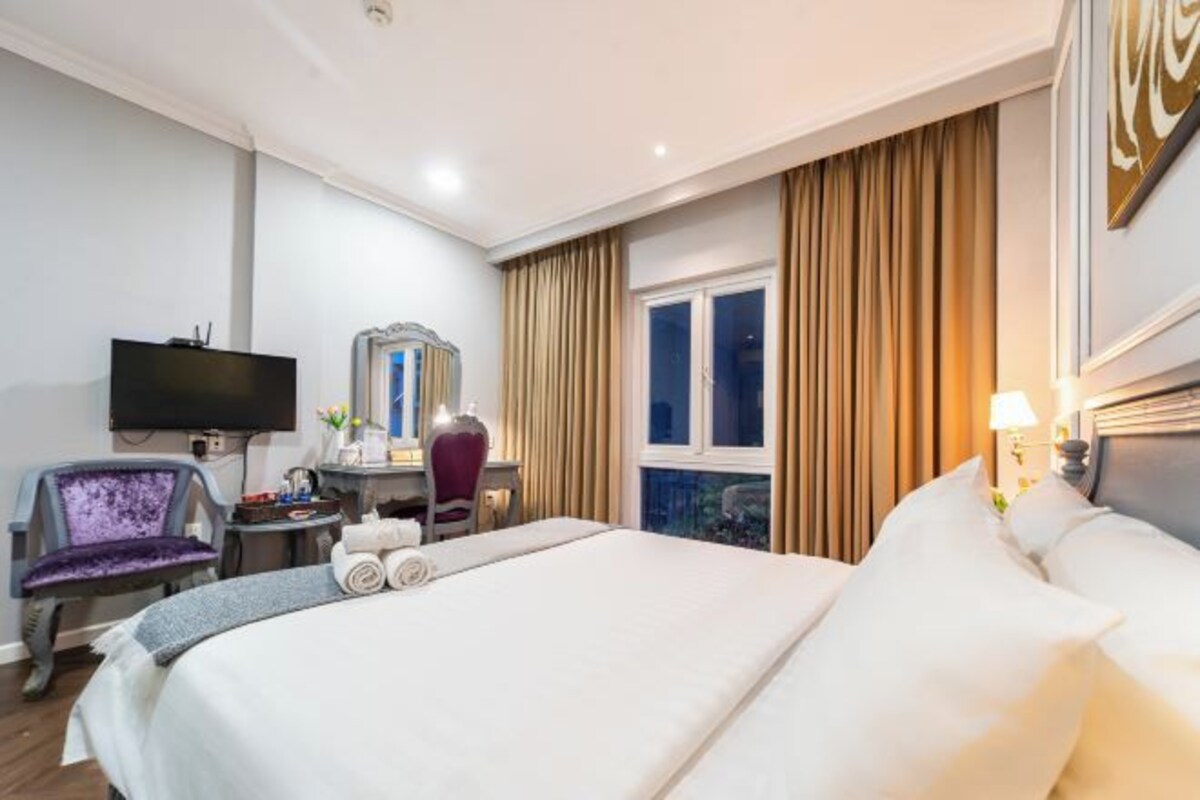 Una habitación de hotel con una cama grande, un escritorio con silla, televisor y ventana con cortinas.