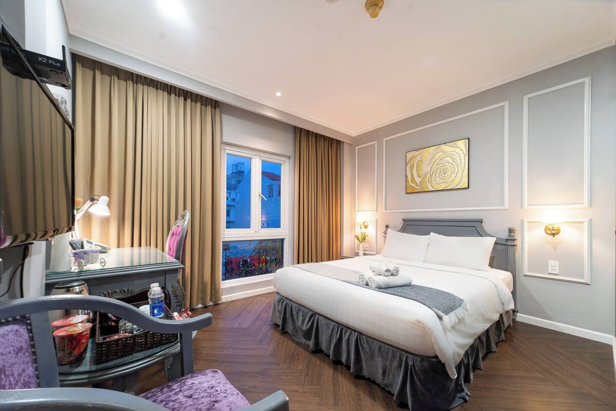 Ein Hotelzimmer mit einem großen Bett, einem Schreibtisch mit Computer, einem Stuhl und einem Fenster mit Vorhängen.