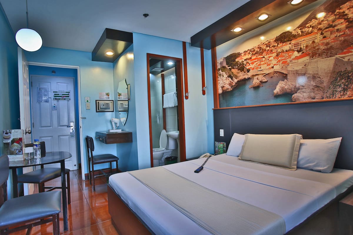 A hotel room with a bed, a desk with a chair, a bathroom, and a wall-mounted TV displaying a scenic view.