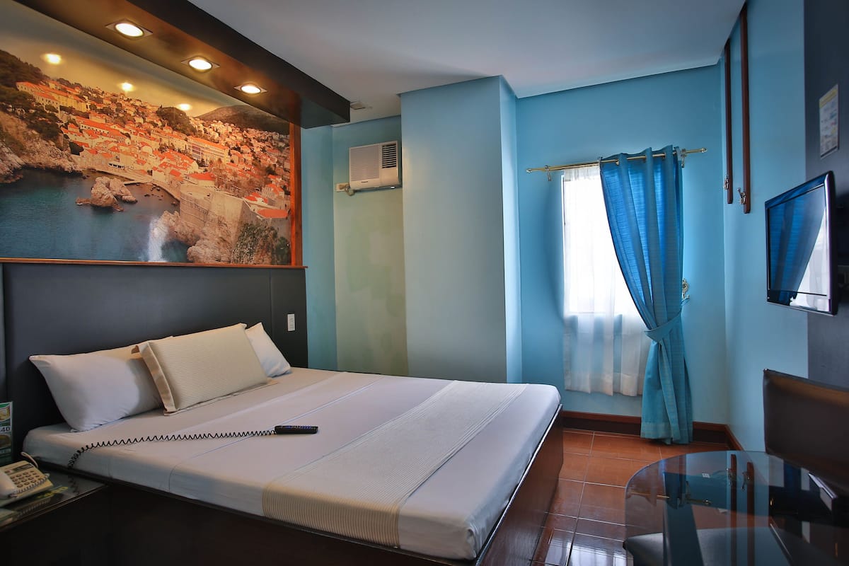 A hotel room with a bed, a TV, a window with curtains, and a wall-mounted air conditioner.