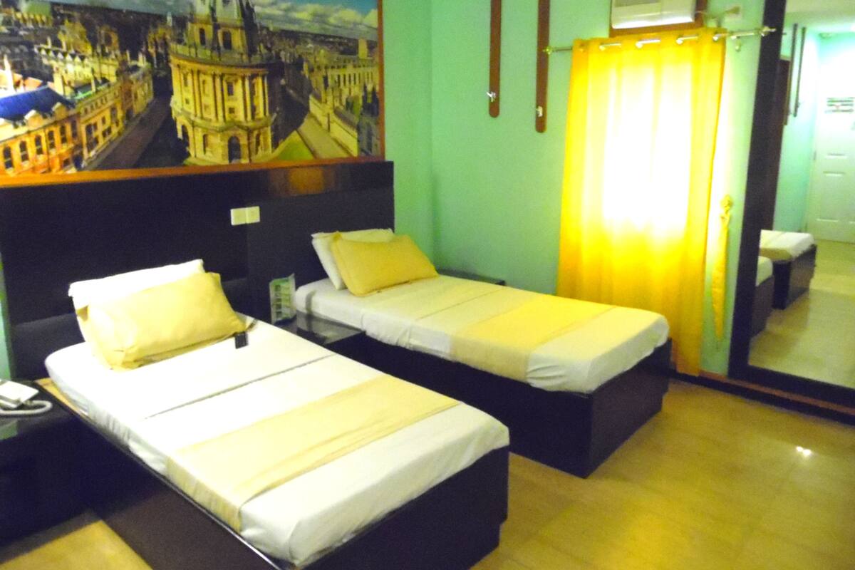 A hotel room with two single beds, a nightstand, a telephone, a painting of a cityscape, a window with curtains, and a mirror on the wall.