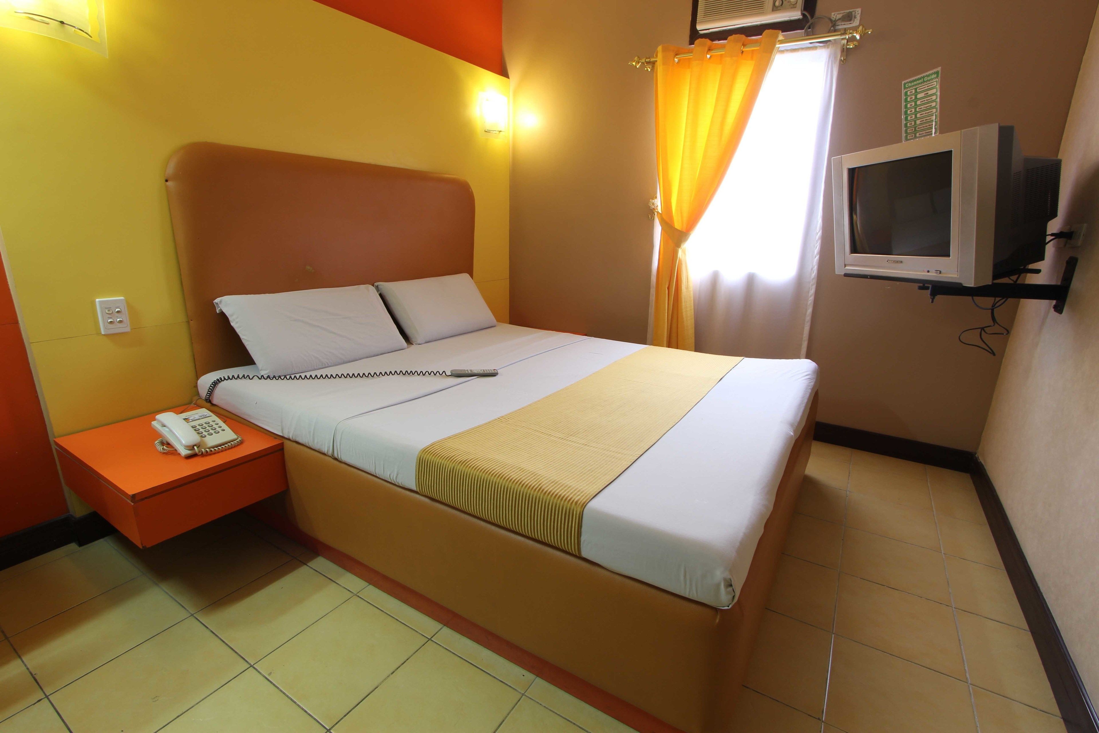 Une chambre d’hôtel avec un lit, une table de chevet avec un téléphone, un téléviseur posé sur un meuble et une fenêtre avec des rideaux.