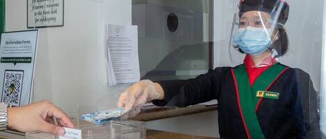 A hotel reception area with a person wearing a uniform and a face shield, handling a card through a transparent barrier.