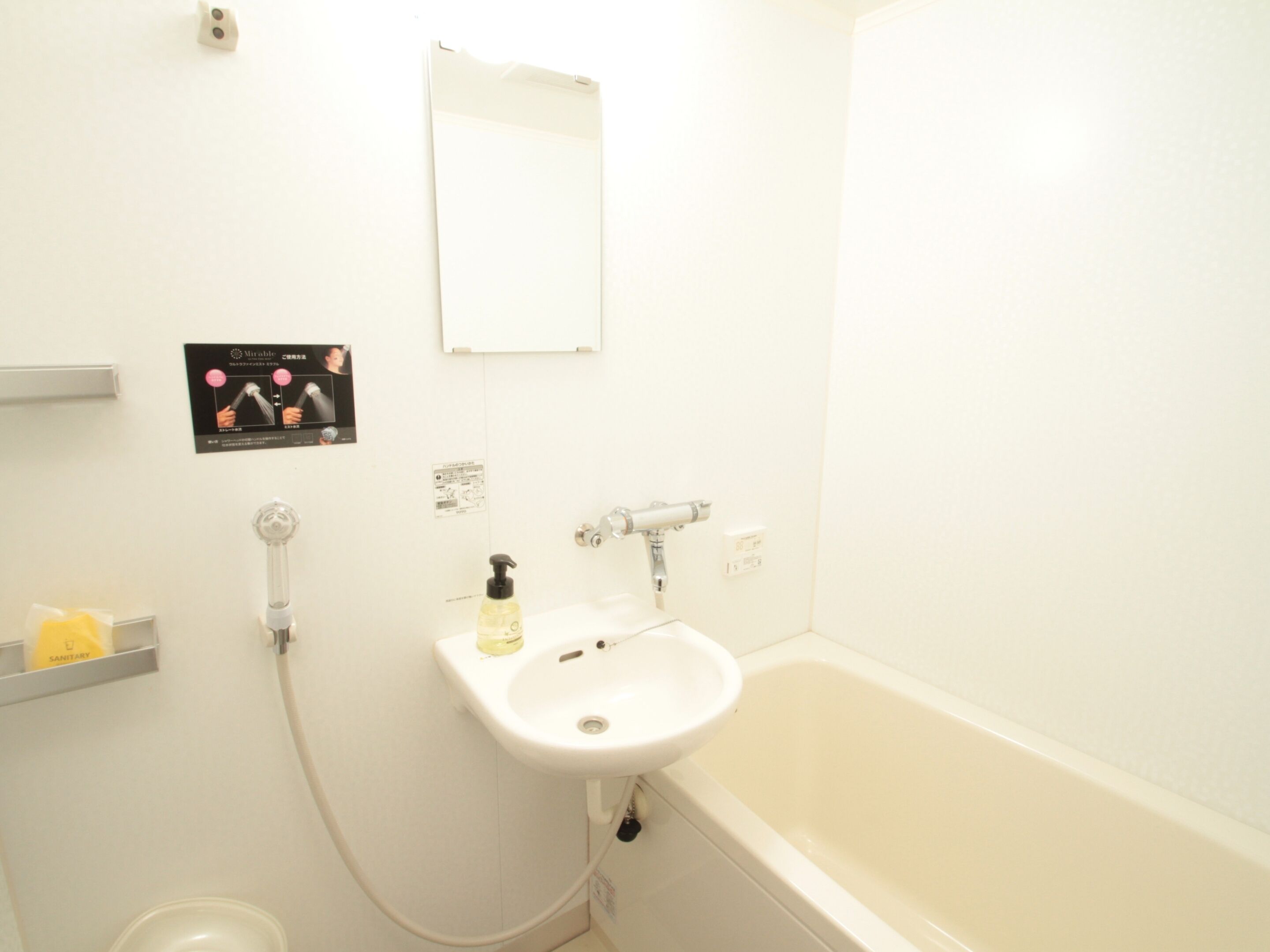 Une salle de bain avec un lavabo, un miroir et une baignoire. On y trouve un distributeur de savon liquide et une petite étagère avec une bouteille jaune.