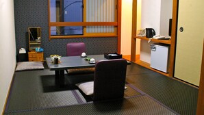 Habitación de hotel compacta con suelo de tatami, una pequeña mesa con una silla, un escritorio con una silla, una ventana con persianas, un espejo, un refrigerador y una estantería con una tetera y un teléfono.