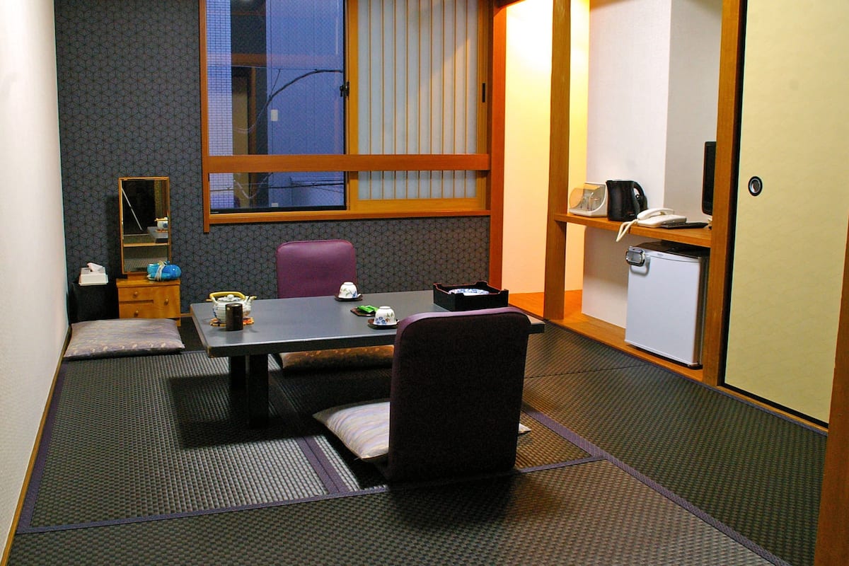 A compact hotel room with a tatami mat floor, a small table with a chair, a desk with a chair, a window with blinds, a mirror, a refrigerator, and a shelf with a kettle and a phone.